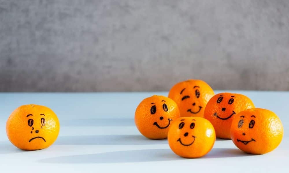 Yalnız bırakılmış, üzgün bir portakalın, gülen diğer portakal grubundan uzakta durduğu, akran zorbalığı veya dışlanma temasını anlatan görsel.